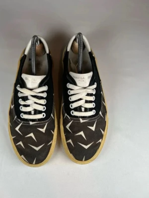 Saint Laurent mönstrade låga sneakers - Snygga låga sneakers från Saint Laurent med svart och beige tyg, vita detaljer och grafiskt mönster. Skorna har vita snören, beige sula och vit hälflik. Perfekta för dig som gillar exklusiv streetstyle och vill sticka ut med ett unikt mönster. Små täcken på användning och ny pris 6500 kr!