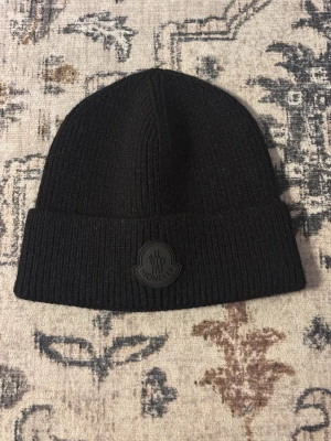 Moncler mössa  - Säljer en svart ribbstickad mössa från Moncler med uppvikt kant och Moncler-logga framtill. Mössan är tillverkad i mjuk ull och har en klassisk, stilren design som passar perfekt till höst och vinter.