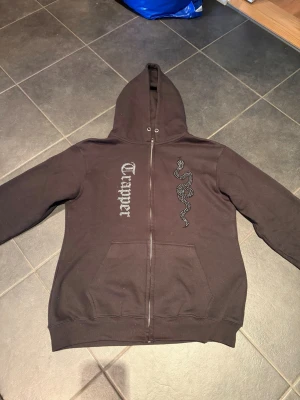 Svart Trapper hoodie - Säljer en svart Trapper hoodie med dragkedja och huva. På framsidan finns en stor orm i glittrande stenar och texten 'Trapper'. Perfekt för dig som gillar streetwear och unika detaljer.