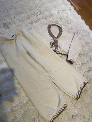 Beige loungewear-set med bruna detaljer - Mysigt loungewear-set i beige med bruna ribbade kanter. Setet består av ett par vida byxor och en långärmad kofta med knappar framtill. Perfekt för chill dagar hemma. Materialet är mjukt stickat tyg som känns skönt mot huden.