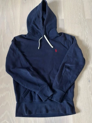 Mörkblå hoodie från Polo Ralph Lauren - Snygg mörkblå hoodie från Polo Ralph Lauren med klassisk röd logga broderad på bröstet. Hoodien har vit dragsko, känguruficka och mjukt material som känns skönt mot huden. Perfekt för en avslappnad och stilren look. Pris går att diskutera. Vid intresse är det bara att höra av sig 