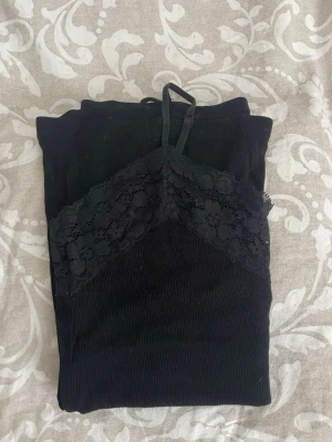 Svart ribbad linnetopp med spets - Snygg svart ribbad linnetopp med tunna axelband och spetsdetaljer vid urringningen. Ena bandet lossnade så jag knöt det, därpå priset.
