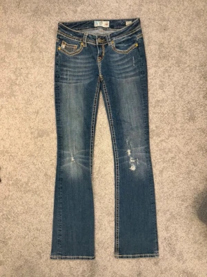 Blå bootcut jeans  - Snygga blå bootcut jeans från Mek Denim med slitningar och broderade detaljer på bakfickorna. Jeansen har kontrastsömmar, låg midja och klassisk femficksmodell. Materialet är denim med stretch för extra komfort.