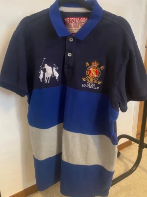 Randig Club Marseille polo t-shirt - Säljer en randig polo t-shirt från Club Marseille med broderade detaljer på bröstet och klassisk krage. T-shirten har breda blå och grå ränder, korta ärmar och två knappar framtill. Perfekt för dig som gillar sportig och stilren look.