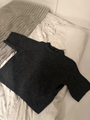 Mörkgrå stickad tröja med halvpolokrage - Mysig mörkgrå stickad tröja med halvpolokrage och kortare ärmar. Perfekt för lager-på-lager och enkel att matcha med jeans eller kjol. Tröjan har en rak passform och är tillverkad i ett mjukt material som känns skönt mot huden. Från lager 157 använd fåtal gånger.