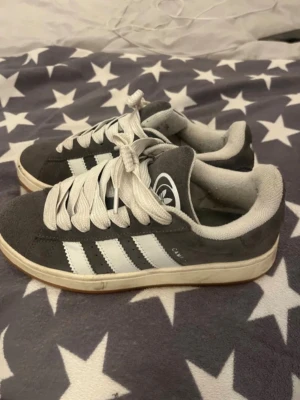 Grå Adidas sneakers med vita ränder - Säljer ett par grå Adidas sneakers med klassiska vita ränder på sidan och vit sula. Skorna har breda vita skosnören och är låga i modellen. Ovandelen är i mocka och insidan är fodrad i vitt tyg. Perfekta för en avslappnad streetstyle. Betalning genom vanlig swish! 