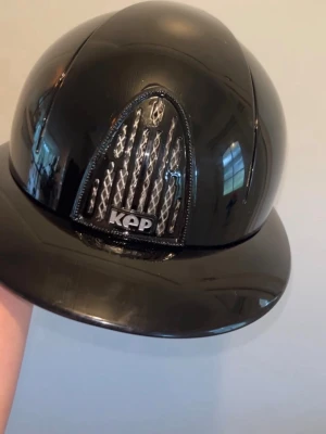 Svart ridhjälm från KEP - Snygg svart ridhjälm från KEP med blank finish och ventiler i mesh framtill. Hjälmen har en klassisk form och en bred skärm som ger extra skydd. Perfekt för dig som vill ha både stil och funktion i stallet.