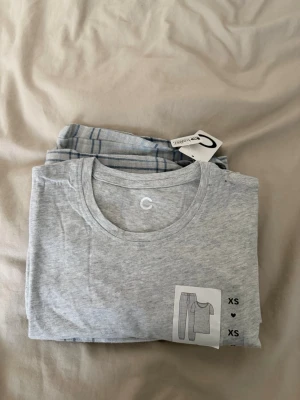 Gråmelerad pyjamas med rutiga byxor - Mysig pyjamas från Cubus i storlek XS. Setet består av en gråmelerad t-shirt med rund hals och ett par ljusgrå byxor med blått rutmönster och dragsko i midjan. Perfekt för chillkvällar hemma. Materialet känns mjukt och bekvämt mot huden.