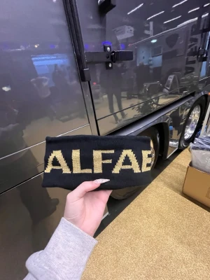 ALFAB - Snyggt svart stickat pannband från ALFAB med stor gul text framtill. Perfekt accessoar för att hålla öronen varma och samtidigt se cool ut. Materialet är mjukt och stretchigt, vilket gör att det sitter skönt på huvudet.