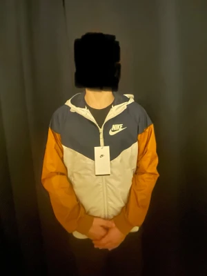 Nike vindjacka - Helt ny Nike vindjacka med prislapp.  Jackan har marinblå axelparti, vita paneler och ärmar i rostbrunt. Utrustad med dragkedja, två sidofickor och resår vid ärmslut och midja. Tillverkad i lätt, vindtätt material. Perfekt för en sportig och trendig look.