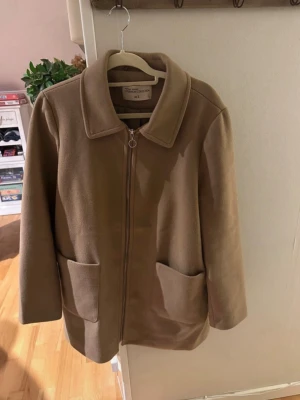 Beige rock från Outerwear Collection storlek L - Stilren beige rock med dragkedja från Outerwear Collection. Klassisk krage, två stora framfickor och rak passform. Tillverkad i mjukt material som ger en clean look. Perfekt för dig som gillar minimalistisk stil.