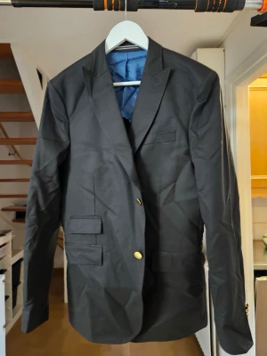 Svart klassisk kavaj med guldknappar - Stilren svart kavaj med två guldknappar framtill och flera fickor med lock. Kavajen har klassisk krage och är helfodrad med blått innerfoder. Passar perfekt till en dressad look och har en tidlös design.pris kan diskuteras!! Storlek M/L