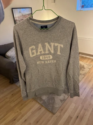 Grå sweatshirt från GANT - Grå sweatshirt från GANT med trycket 'GANT 1949 NEW HAVEN' på bröstet. Tröjan har rund hals, ribbade muddar vid ärmslut och nederkant samt en avslappnad passform. Tillverkad i mjukt bomullsmaterial.