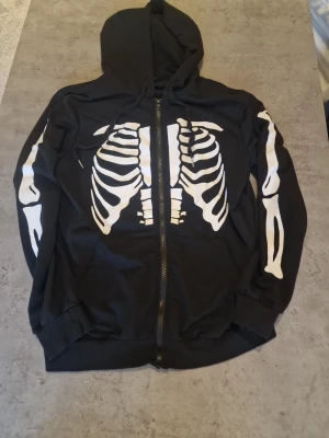 Svart hoodie med skelettmönster - Cool svart hoodie från Bik Bok med dragkedja och vitt skelettmönster på både framsidan och ärmarna. Hoodien har huva med dragsko och ribbade muddar. Perfekt för dig som gillar edgy y2k och unika plagg. Eller bara en hoodie till halloween