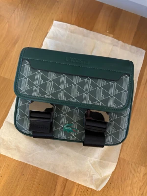 Grön mönstrad handväska från Lacoste - Snygg grön handväska från Lacoste med grafiskt mönster i vitt och två svarta spännen framtill. Väskan har ett klassiskt Lacoste-märke med krokodil på framsidan och är gjord i tåligt konstläder. Perfekt för dig som vill ha en unik accessoar med sportig touch.