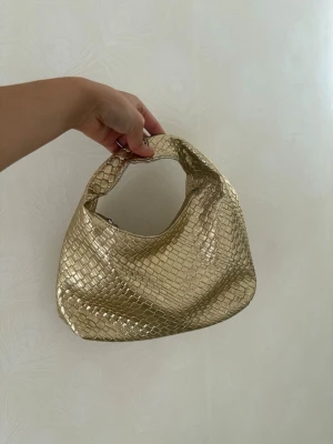 Guldig flätad handväska - Trendig guldig handväska i flätad look med mjuk, rundad form och kort handtag. Väskan har dragkedja upptill och skimrande metallic-finish som ger en lyxig känsla. Perfekt accessoar för att lyfta vilken outfit som helst.