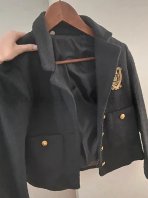 Svart kavaj med guldknappar och emblem - Stilren svart kavaj med guldknappar och två framfickor. Kavajen har ett broderat guldemblem på bröstet och är fodrad med svart tyg. Klassisk krage och rak passform ger en tidlös look. Perfekt för dig som gillar preppy och elegant stil.
