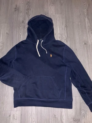 Mörkblå hoodie från Polo Ralph Lauren - Mörkblå hoodie från Polo Ralph Lauren med klassisk huva och vit dragsko. Har en broderad gul logga på bröstet och en stor känguruficka framtill. Tillverkad i mjukt bomullsmaterial, perfekt för en avslappnad stil.