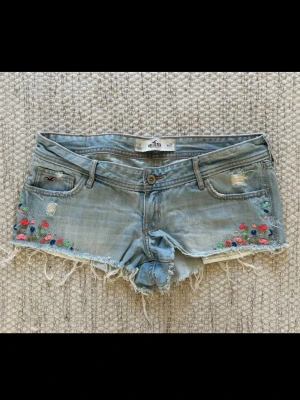 Broderade jeansshorts från Hollister - Snygga ljusblå jeansshorts från Hollister med slitna detaljer, fransig kant och broderade blommor i rött, grönt och blått på framsidan. 