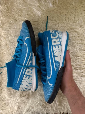 Nike Mercurial blå fotbollsskor - Säljer ett par Nike Mercurial fotbollsskor i blått med vita detaljer och svart sula. Skorna har högt skaft, snörning och stor vit Nike-logga på sidan. Ovandelen är i mesh-liknande material för bra ventilation och lätt känsla på planen.