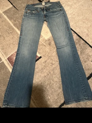 Vintage jeans från True Religion - Säljer ett par blå bootcut jeans från True Religion med klassiska kontrastsömmar och snygga detaljer på bakfickorna. Jeansen har låg midja och är tillverkade i mjukt denimtyg. Perfekta för dig som gillar en avslappnad men trendig stil.