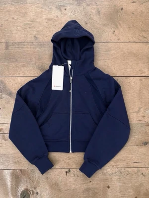 Mörkblå hoodie från Lululemon - Snygg mörkblå hoodie från Lululemon med dragkedja framtill, huva och två fickor. Croppad modell med breda muddar vid ärmslut och nederkant. Perfekt för en sportig och avslappnad stil. Materialet är mjukt och stretchigt för extra komfort.                                                                                                                                                        storlek xs