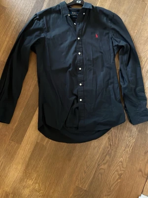 Svart skjorta från Polo Ralph Lauren - Klassisk svart skjorta från Polo Ralph Lauren med lång ärm och knappar framtill. Skjortan har den ikoniska röda logotypen broderad på bröstet och är tillverkad i mjuk bomull. Perfekt för en clean och stilren look. Skjortan är äkta utan tecken på användning. Tveka inte på att höra av er vid frågor