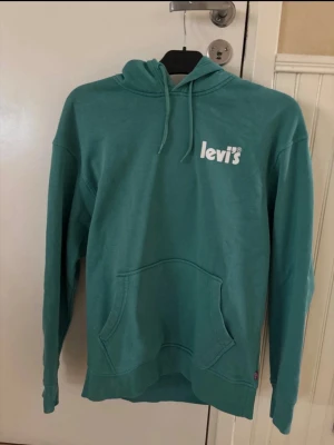 Turkos hoodie från Levi's - Snygg turkos hoodie från Levi's med vit logga på bröstet. Klassisk känguruficka framtill, justerbar huva med dragsko och ribbade muddar vid ärmslut och nederkant. Perfekt för en avslappnad och trendig stil.