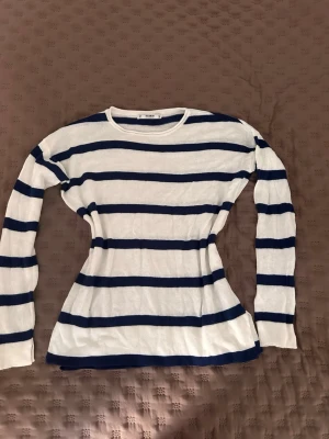 Randig stickad tröja från Pull&Bear - Tunt stickad tröja från Pull&Bear i vitt med mörkblå horisontella ränder. Tröjan har rund halsringning och långa ärmar. Perfekt för en avslappnad och stilren look. Materialet är mjukt och lätt, vilket gör den skön att bära.