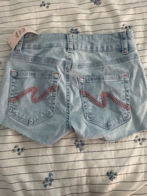 Ljusblå jeansshorts från Nelly - Snygga ljusblå jeansshorts från Nelly med råa kanter och dubbla knappar framtill. Shortsen har bakfickor med rosa broderad vågdetalj och klassisk femficksdesign. Perfekta för varma dagar och enkel att matcha med allt.