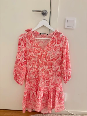 Rosa och vit mönstrad klänning - Söt klänning med rosa och vitt blommönster, v-ringning och trekvartsärmar med resår. Klänningen har volangkant nertill och en liten krage. Perfekt för en somrig look.