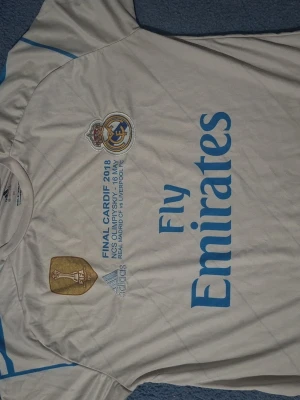 Real Madrid Ronaldo matchtröja 2018 - Vit Real Madrid matchtröja från Adidas med blå detaljer, tryck 'Fly Emirates' och FIFA-märke. Final Cardiff 2018-tryck på bröstet och Ronaldo 7 på ryggen. Perfekt för fotbollsfans som vill sticka ut på planen eller läktaren.