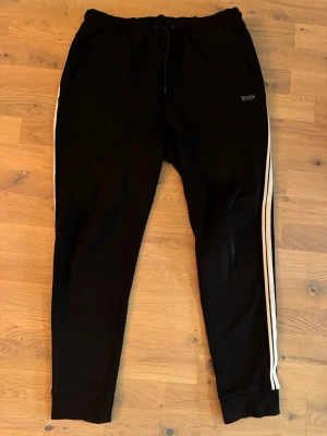 Svarta mjukisbyxor från BOSS - Svarta mjukisbyxor från BOSS med vita sidoränder och dragsko i midjan. Byxorna har smal passform och ribbade muddar vid bensluten. Perfekta för en avslappnad och sportig stil.