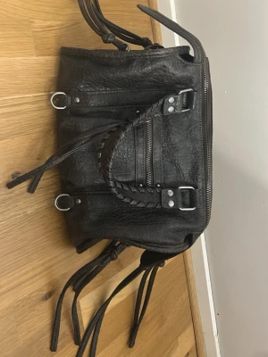 Svart handväska med flätat handtag - Cool svart handväska i skinn med flätat handtag, silverfärgade metalldetaljer och dragkedja framtill. Väskan har flera dekorativa remmar och ringar samt en praktisk ytterficka med dragkedja. Perfekt för dig som gillar en edgy och trendig look.