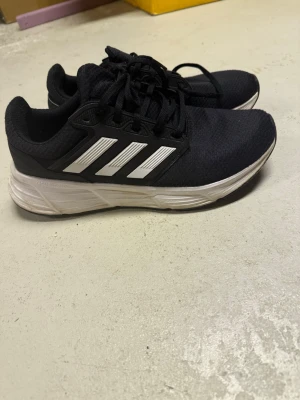 Svarta Adidas sneakers med vita ränder - Svarta sneakers från Adidas med klassiska vita ränder på sidorna och vit sula. Ovandelen är i meshmaterial för bra andningsförmåga och skon har snörning framtill. Perfekt för en sportig och avslappnad stil. Se bilder 