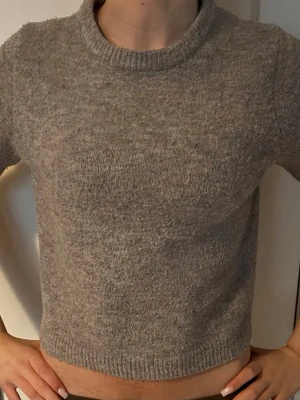 Grå stickad kortärmad tröja - Mysig grå stickad tröja med kortare passform och rund halsringning. Tröjan har ribbade muddar vid hals och nederkant, och är perfekt för lager-på-lager-stil. Enkel och stilren design som passar till det mesta.