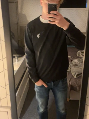 Svart crewneck från U.S. Polo Assn. - Svart sweatshirt med rund hals från U.S. Polo Assn. Tröjan har en vit broderad logga på bröstet och ribbade muddar vid ärmslut och nederkant. Perfekt för en avslappnad och stilren look.