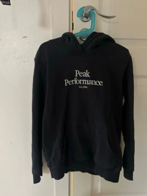 Mörkblå hoodie från Peak Performance - Mörkblå hoodie med vit Peak Performance-logga på bröstet. Klassisk modell med huva och känguruficka framtill. Perfekt för en avslappnad och sportig stil. Mjuk bomullsblandning och ribbade muddar vid ärmslut och nederkant.