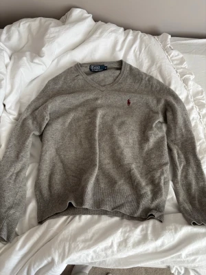 Grå v-ringad tröja från Ralph Lauren - Klassisk grå v-ringad tröja från Ralph Lauren med broderad röd logga på bröstet. Tröjan har långa ärmar och ribbade muddar vid ärmslut och nederkant. Perfekt att slänga på sig över en skjorta eller t-shirt.