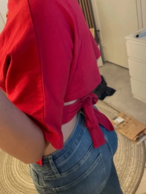 Röd topp med knyt i ryggen - Snygg röd  topp med korta ärmar och knyt i ryggen. Perfekt till jeans eller kjol. Tillverkad i mjuk bomull som känns skön mot huden. Enkel men ändå unik detalj med knytband bak. Endast använd en gång