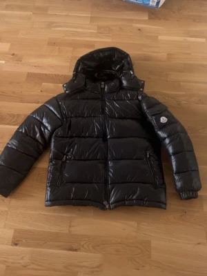 Svart dunjacka från Moncler - Snygg svart dunjacka från Moncler med glansig finish, huva och två dragkedjefickor. Klassisk Moncler-logga på ärmen och robust dragkedja framtill. Perfekt för kalla vinterdagar och riktigt trendig look.