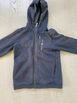 Mörkblå hoodie från Sail Racing strl 160 - Mörkblå hoodie med dragkedja från Sail Racing. Tröjan har huva med dragsko, bröstficka med dragkedja och två sidofickor. Mjuk bomullskänsla och stilren design med logga på bröstet. Perfekt för en avslappnad och sportig look.