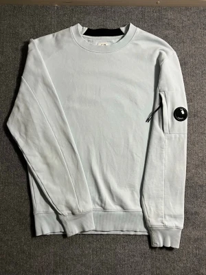 C.P. Company – Turquoise Lens Sweatshirt (M) - Fresh and stylish C.P. Company sweatshirt in a vibrant turquoise shade. Size M, length 65 cm, width 50 cm – designed for a modern, relaxed fit. The sleeve features the signature lens detail, giving this piece its unmistakable character and premium edge.  ✅ Condition: Excellent – no signs of wear or stains 💯 Authenticity: 100% genuine, easily verified on certilogo.com 📦 Shipping: Dispatched immediately after purchase – fast and smooth transaction!  This sweatshirt blends C.P. Company’s technical 