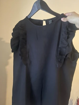 Svart volangblus från Shein - Säljer en svart blus från Shein med volangdetaljer längs axlarna. Toppen är ärmlös och har en rund halsringning samt knapp i nacken. Perfekt för dig som gillar stilrena plagg med en twist.
