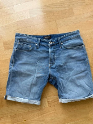 Ljusblå jeansshorts från Jack & Jones - Snygga ljusblå jeansshorts från Jack & Jones med klassisk femficksdesign och uppvikta benslut. Shortsen har normal passform och bälteshällor i midjan. Perfekta för varma dagar och enkla att matcha med olika stilar.