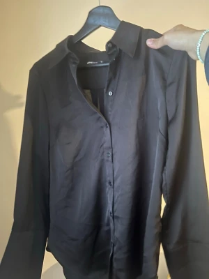 Svart skjorta från Gina Tricot - Stilren svart skjorta från Gina Tricot med klassisk krage och knäppning framtill. Skjortan har lång ärm och är tillverkad i ett lätt, något glansigt tyg som ger en snygg look. Perfekt att styla med jeans eller kjol.