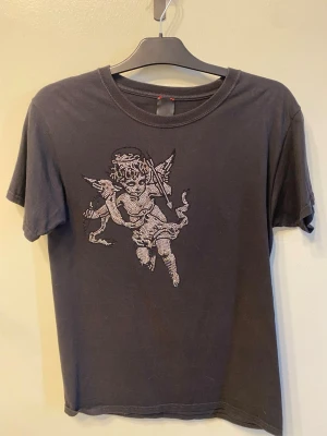 Svart t-shirt med ängeltryck - En snygg svart bvaldi tröja med en ängel på framsidan. T-shirten har rund hals och korta ärmar. Perfekt för dig som gillar streetwear och unika prints. Tröjan har tappat lite diamanter både där fram och bak men på framsidan märker man inte det om man inte kollar noga. Köpt på bvaldis affär i Helsingborg på väla. 