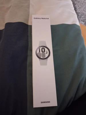 Samsung Galaxy Watch4 smartklocka - Samsung Galaxy Watch4 är en stilren smartklocka med rund urtavla och vit silikonrem. Klockan har en modern design och levereras i originalförpackning. USB-laddkabel ingår. Perfekt för dig som vill ha koll på hälsa, träning och notiser direkt på handleden.
