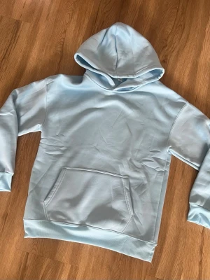 Ljusblå hoodie med magficka - Snygg ljusblå hoodie med klassisk huva och stor magficka framtill. Hoodien har ribbade muddar vid ärmslut och nederkant. Perfekt för en avslappnad och trendig stil. Tillverkad i mjukt material för extra komfort.