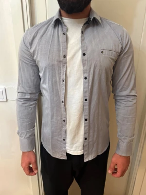 Grå skjorta från Emilio, Small - Snygg grå skjorta från Emilio i storlek Small. Skjortan har lång ärm, klassisk krage, svarta knappar och en bröstficka med knappdetalj. Perfekt att bära öppen över en t-shirt för en avslappnad look.